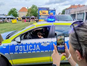 Policyjne stoisko, namiot, ludzie, mierzenie prędkości w biegu, przymierzanie strojów, dzieci uśmiechnięte w radiowozie.