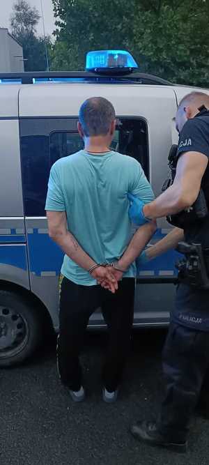 Mężczyzna przed radiowozem, w kajdankach obok policjant